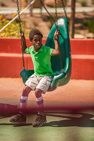 A boy sits on the edge of an accessible green swing, an important element of accessible playground equipment 