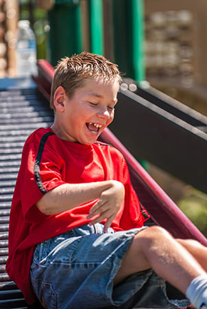 A little boy laughs as he has just finished sliding down the rollerslide, an element of accessible playground equipment. 