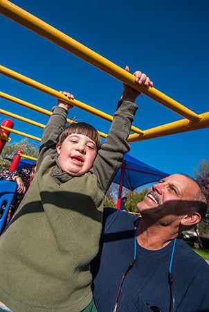 A boy smiles and a man helps him as he navigates an overhead monkeybar structure, an example of wheelchair accessible playground equipment. 