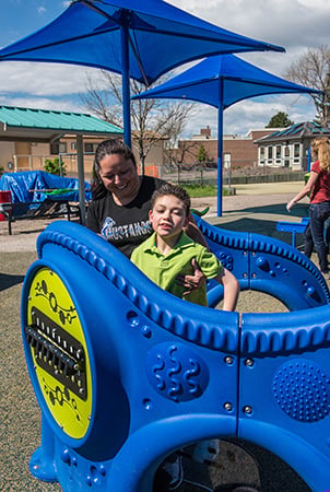 A boy stands inside a wheelchair accessible playground equipment structure while a woman helps him from outside. 