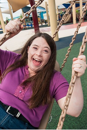 A young woman laughs as she swings sideways on an accessible playground equipment swing. 