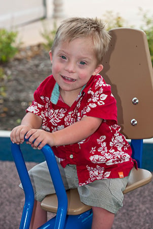 A little boy in a red Hawaiian shirt sits in an adaptive seat, a component of accessible playground equipment. 
