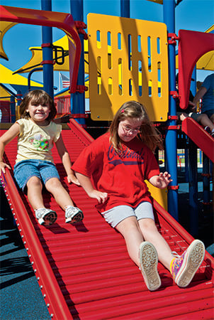 Two girls slide down a red rollerslide, an example of accessible playground equipment. 