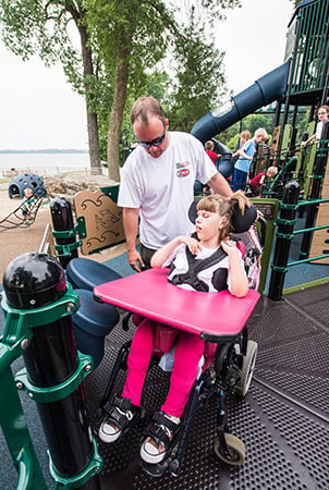 A man and a girl in a wheelchair are on a PlayBooster deck and they are looking at the bongo drum panel, which is a playground accessibility structure. 